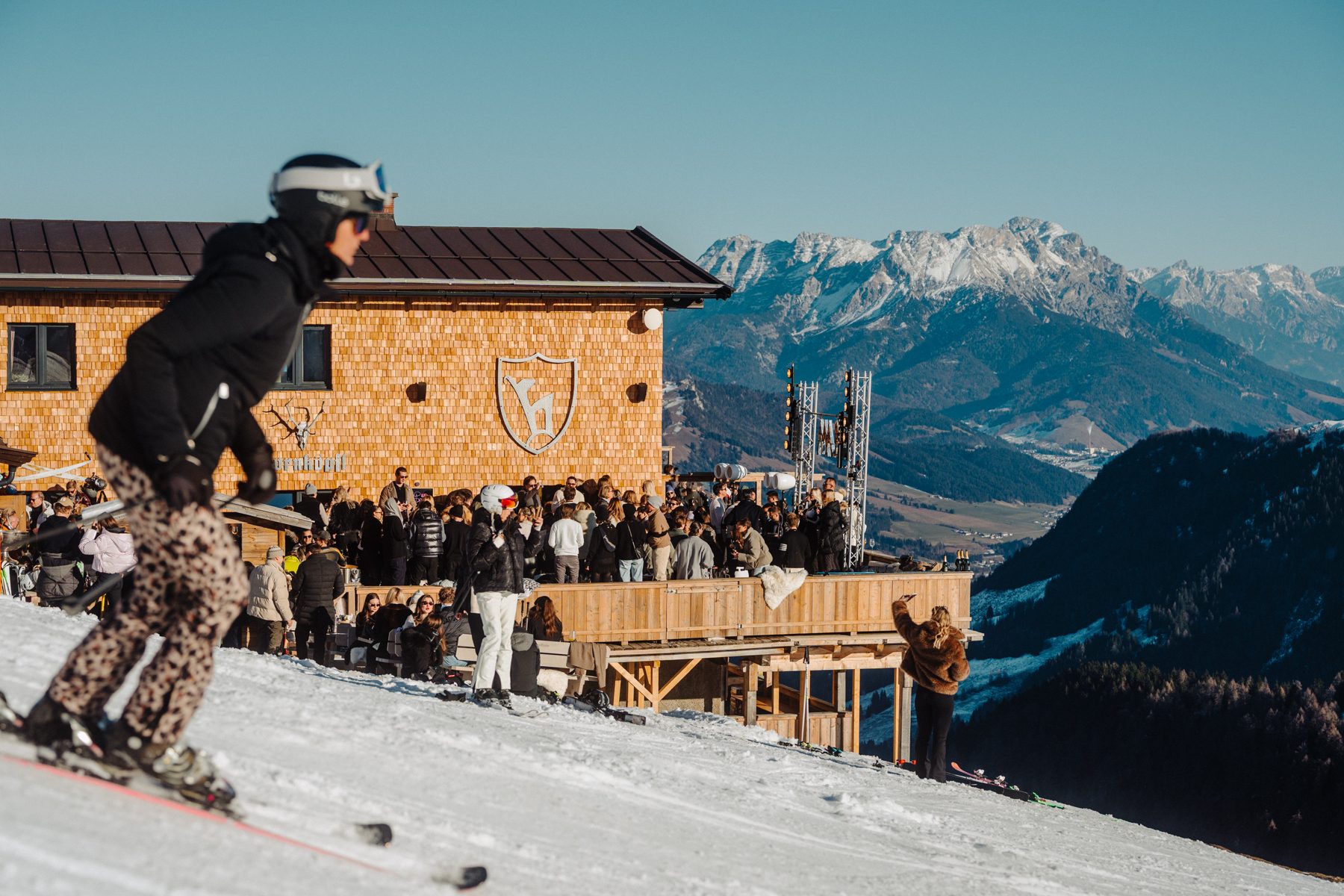 Skifahrerin auf der Piste vor der vollen Après-Ski-Terrasse der Hornköpflhütte